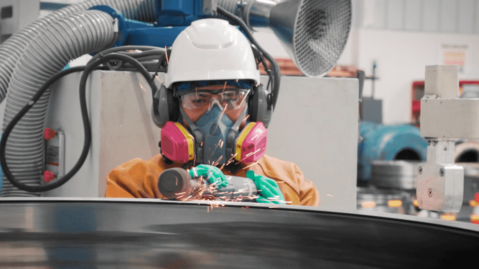 Un trabajador con una camisa naranja, un casco de seguridad blanco, orejeras grises, y máscara protectora, trabaja con un equipo de soldadura sobre una pieza de metal, con chispas volando. Al fondo se ven equipos industriales y maquinaria.